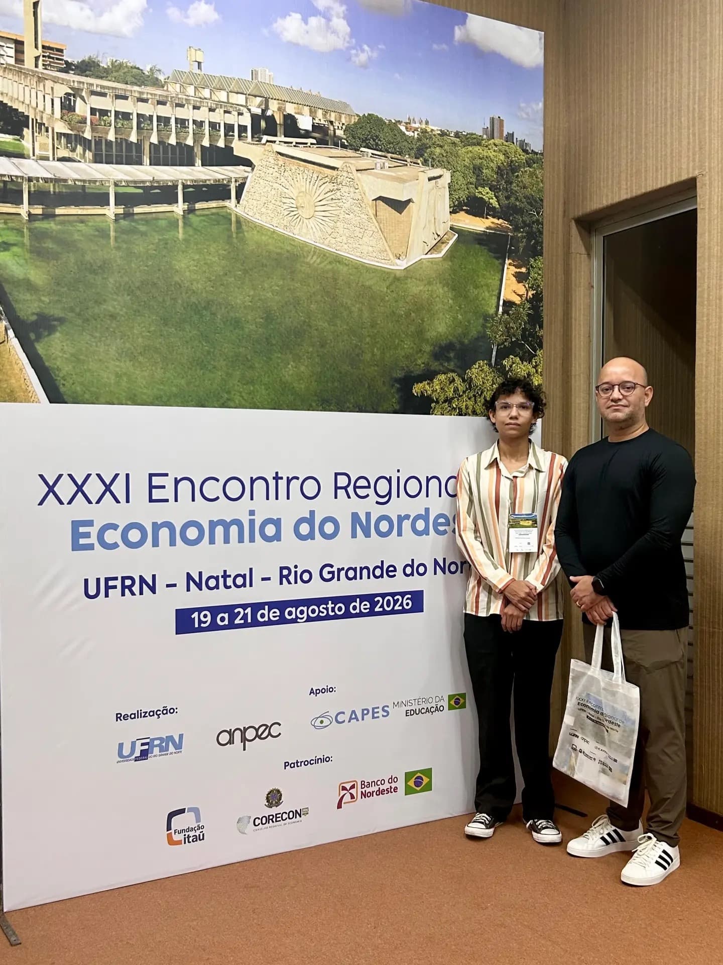 Duas pessoas ao lado do painel do XXXI Encontro Regional de Economia do Nordeste, na UFRN